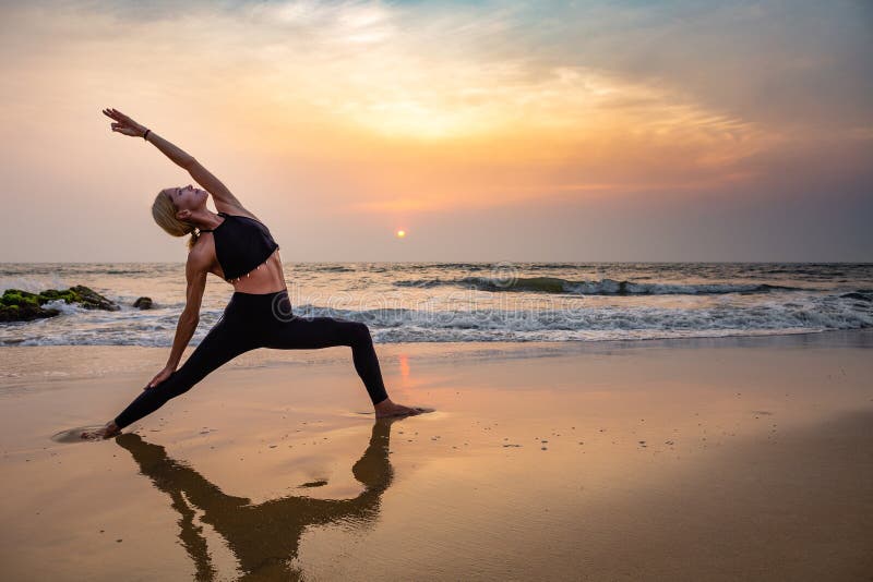 Middle age woman in black doing yoga on sand beach in India  standing asana virabhadrasana at sunset. Healthy lifestyle