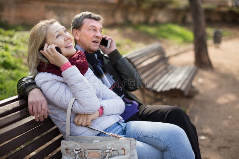 Middle Aged Couple Talking on the Phone on a Bench in the Park Stock