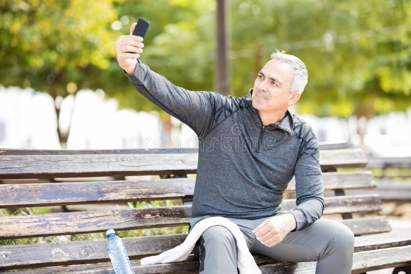 Middle-aged man taking a selfie while sitting in a park bench after exercis...
