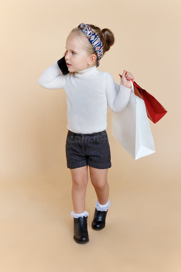 Mignonne petite fille en tenue d'automne tenant à la main un sac de shopping, parlant sur un téléphone portable, sur fond beige photographie stock libre de droits Mignonne petite fille en tenue d'automne tenant à la main un sac de shopping, parlant sur un téléphone portable, sur fond beige photographie stock libre de droits