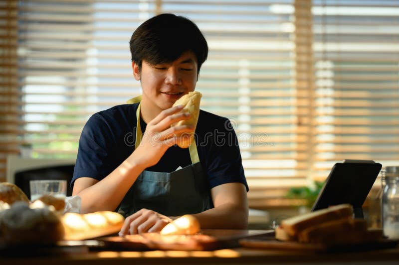 Millennial asiático vestido con ropa informal sosteniendo pan fresco y usando una laptop en mesa de comedor en la cocina.