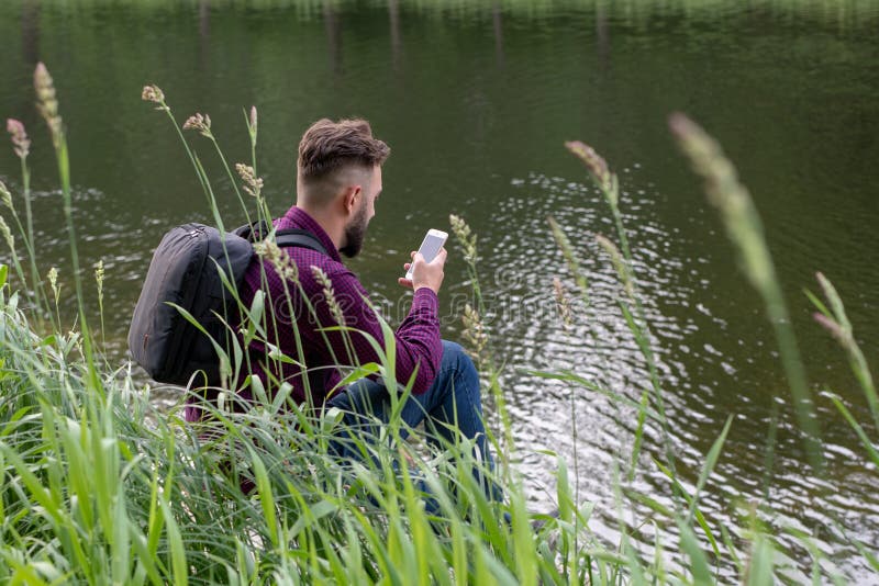 Millennial, młody faceta obsiadanie na plaży, brzeg rzekich spojrzenia w telefon, bierze fotografię Surfingu internet w telefonie zdjęcia stock Millennial, młody faceta obsiadanie na plaży, brzeg rzekich spojrzenia w telefon, bierze fotografię Surfingu internet w telefonie zdjęcia stock