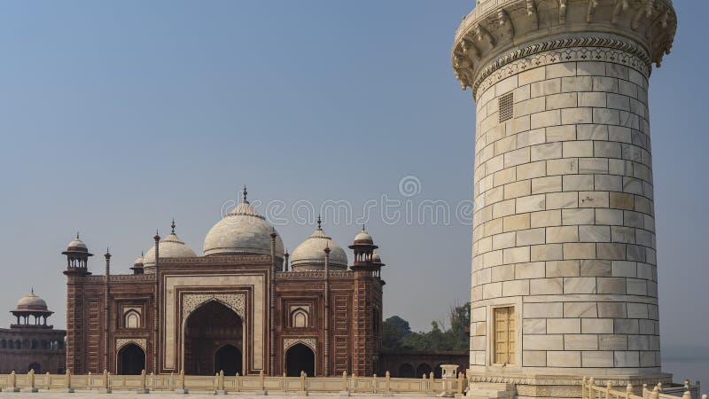 The white marble minaret of the Taj Mahal mausoleum against the blue sky. In the distance, the red sandstone Kau Ban mosque with domes and arches is visible. Blue sky. India. Agra. The white marble minaret of the Taj Mahal mausoleum against the blue sky. In the distance, the red sandstone Kau Ban mosque with domes and arches is visible. Blue sky. India. Agra