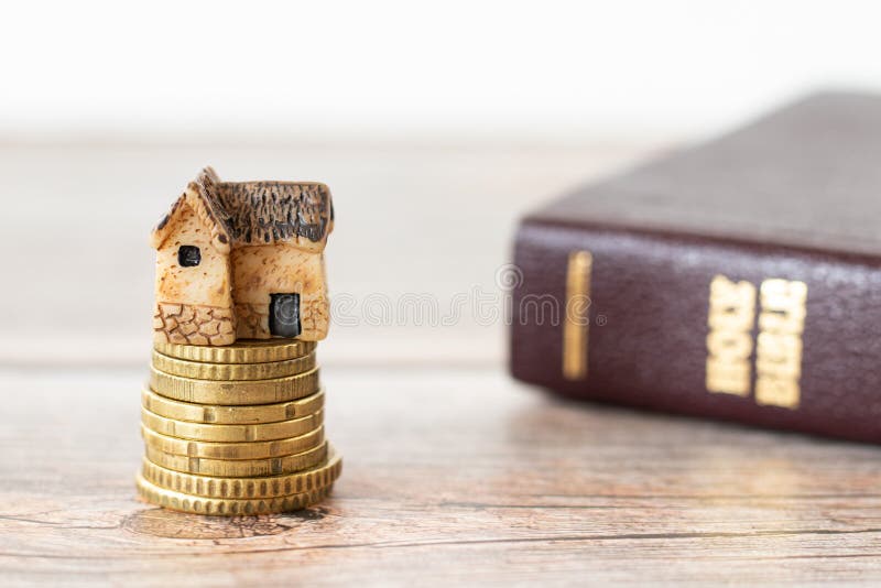 Miniature house on top of a golden stack of coins with a closed Holy Bible Book on a wooden table