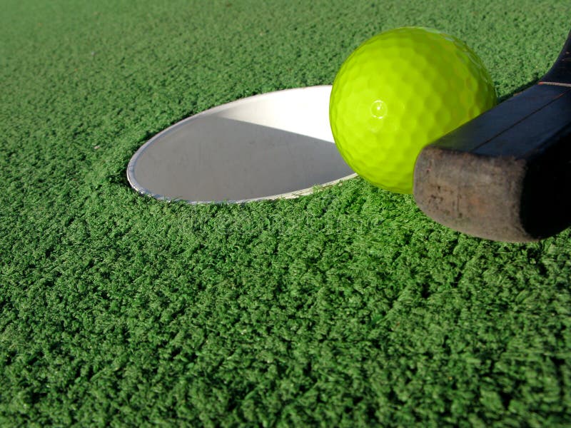 Mini golf club ready to sink the putt with a yellow ball in a hole on artificial green turf at a miniature golfing amusement park. Mini golf club ready to sink the putt with a yellow ball in a hole on artificial green turf at a miniature golfing amusement park