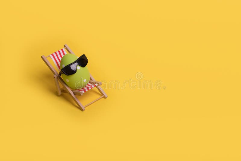 Minimal funny composition with Easter egg with sunglasses while sitting on deck chair on illuminating yellow background royalty free stock photography.