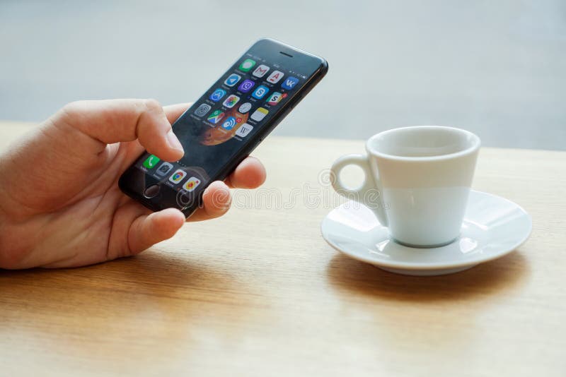 Minsk, Bielorrusia, el 17 de julio de 2017: Mano usando Iphone con los iconos de la aplicación móvil con una taza de café en la t imágenes de archivo libres de regalías Minsk, Bielorrusia, el 17 de julio de 2017: Mano usando Iphone con los iconos de la aplicación móvil con una taza de café en la t imágenes de archivo libres de regalías