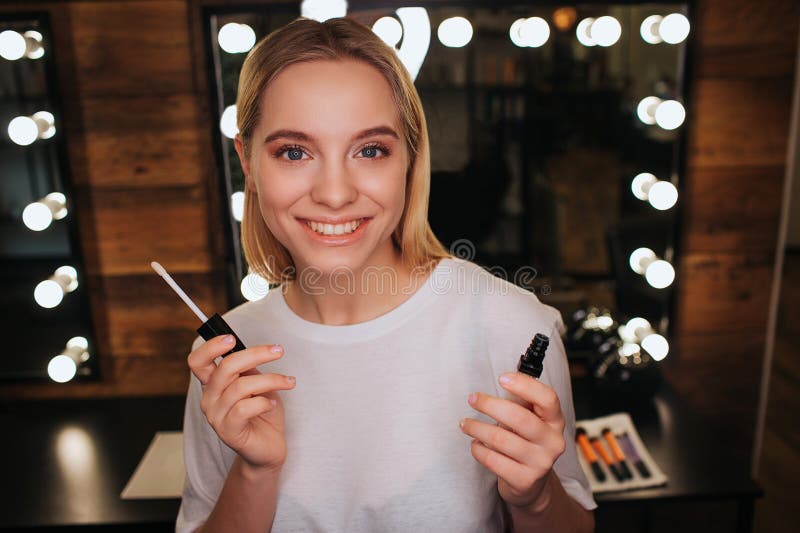 Mirada positiva alegre de la mujer joven en cámara y sonrisa Ella lleva a cabo lipgloss y actitud Espejo con las bombillas detrás imagen de archivo libre de regalías Mirada positiva alegre de la mujer joven en cámara y sonrisa Ella lleva a cabo lipgloss y actitud Espejo con las bombillas detrás imagen de archivo libre de regalías