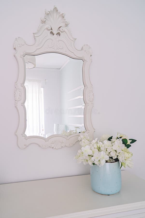 Mirror in carved wooden frame and vase with flowers in bright bedroom interior