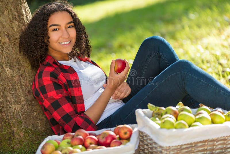 Beautiful happy mixed race African American girl teenager female young woman in an orchard eating an organic red apple, smiling with perfect teeth leaning against a tree with baskets of apples she has been picking. Beautiful happy mixed race African American girl teenager female young woman in an orchard eating an organic red apple, smiling with perfect teeth leaning against a tree with baskets of apples she has been picking