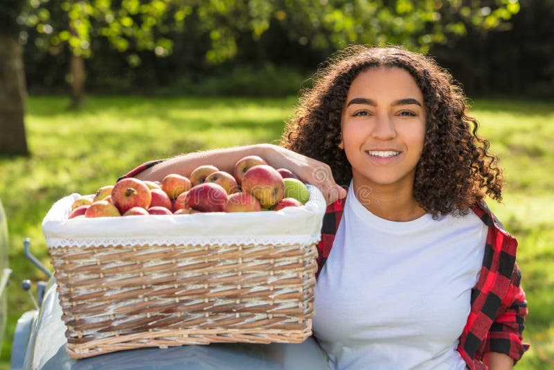 Beautiful happy mixed race African American girl teenager, female young woman, smiling with perfect teeth in an orchard, resting on a tractor with baskets of organic apples she has been picking. Beautiful happy mixed race African American girl teenager, female young woman, smiling with perfect teeth in an orchard, resting on a tractor with baskets of organic apples she has been picking.