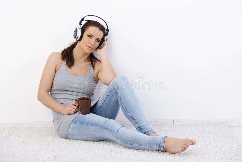 Mid-adult woman enjoying music via headphones, holding coffee mug, sitting on floor, daydreaming. Mid-adult woman enjoying music via headphones, holding coffee mug, sitting on floor, daydreaming.