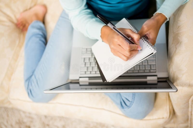 Mittlerer Abschnitt des Frauenschreibens auf Notizblock mit Laptop auf ihren Beinen lizenzfreie stockbilder