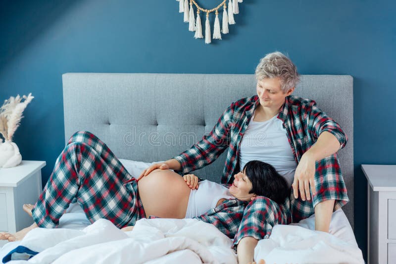 Mixed race couple are waiting for a baby. Husband and his pregnant wife in cozy pajamas relaxing on the bed at modern stock photography