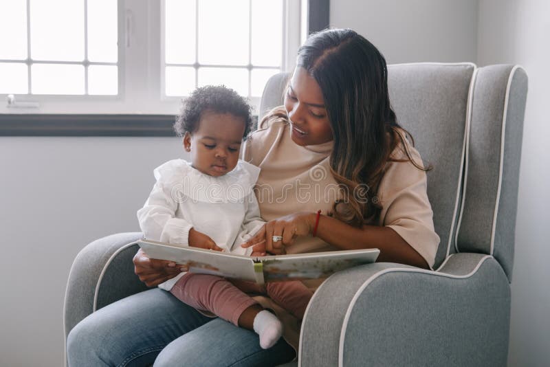 Mixed race Indian mom reading book with African black baby girl toddler at home. Early age children education and development.