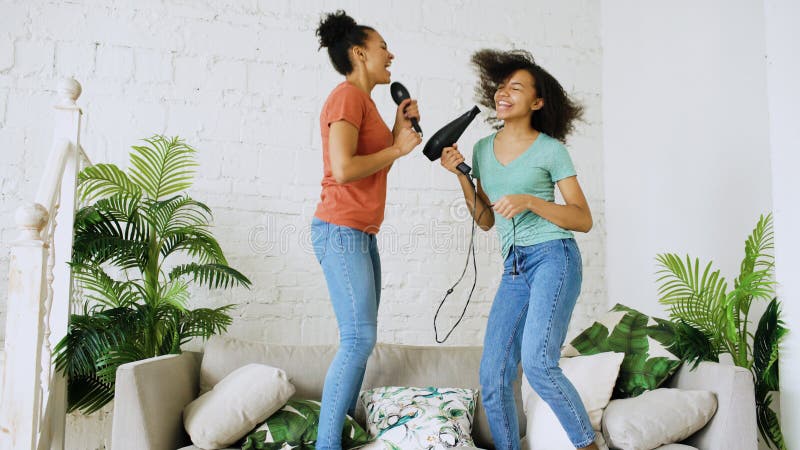 Mixed race young funny girls dance singing with hairdryer and comb jumping on sofa. Sisters having fun leisure in living royalty free stock images Mixed race young funny girls dance singing with hairdryer and comb jumping on sofa. Sisters having fun leisure in living royalty free stock images