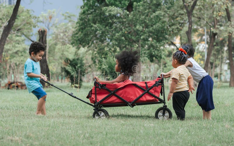 Mixte race petits enfants mignons s'aidant les uns les autres à pousser et traîner un chariot rouge tout en jouant dans le parc ve