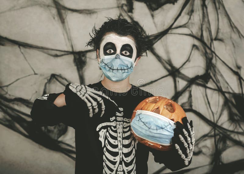 Miúdo feliz do halloween a usar máscara médica numa fantasia esquelética com abóbora do halloween