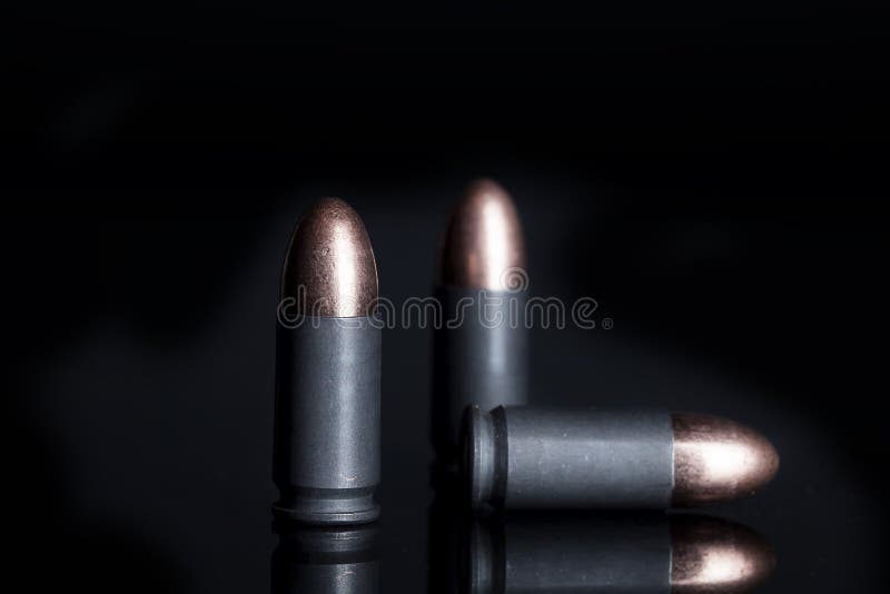 Three 9mm steel cartridges of ammunition, two standing on end and one laying sideways on black reflective surface. Three 9mm steel cartridges of ammunition, two standing on end and one laying sideways on black reflective surface.