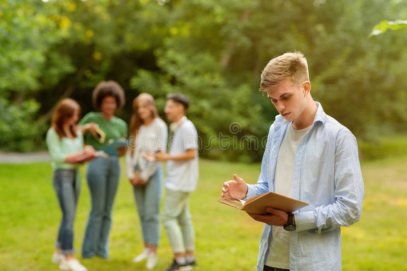 College Bullying. Lonely student guy standing with book at campus apart from classmates, reading, preparing for exam alone, selective focus. College Bullying. Lonely student guy standing with book at campus apart from classmates, reading, preparing for exam alone, selective focus