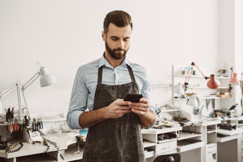 Mobiele toepassing voor het werk Sluit omhoog portret van jonge mannelijke juwelier in schort die een smartphone houden en in mob stock foto Mobiele toepassing voor het werk Sluit omhoog portret van jonge mannelijke juwelier in schort die een smartphone houden en in mob stock foto