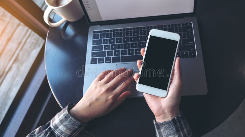 Mockup image of business woman holding mobile phone with blank black screen while using laptop on table stock photo