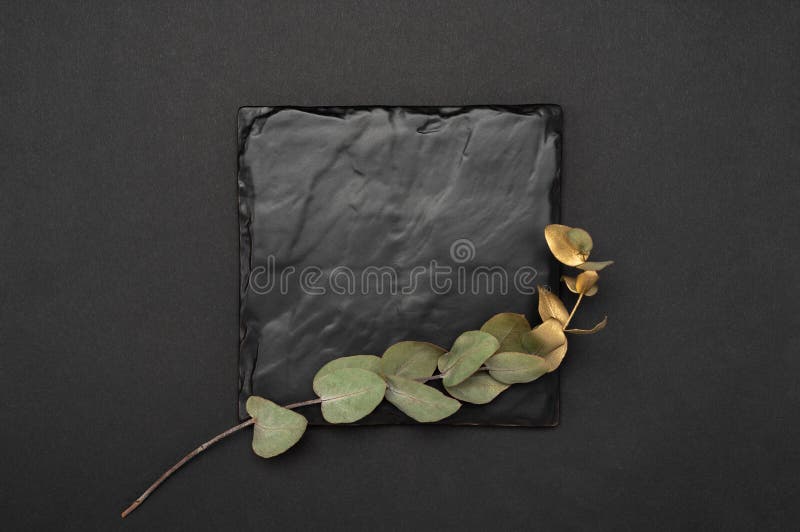 Mockup image. Square black stone plaque plate mock up with a gold eucalyptus branch. Top view of a black card mockup with branch. Square invitation card mock up with a golden eucalyptus branch. Top view of black stone plaque mockup with branch of eucalyptus on black table.