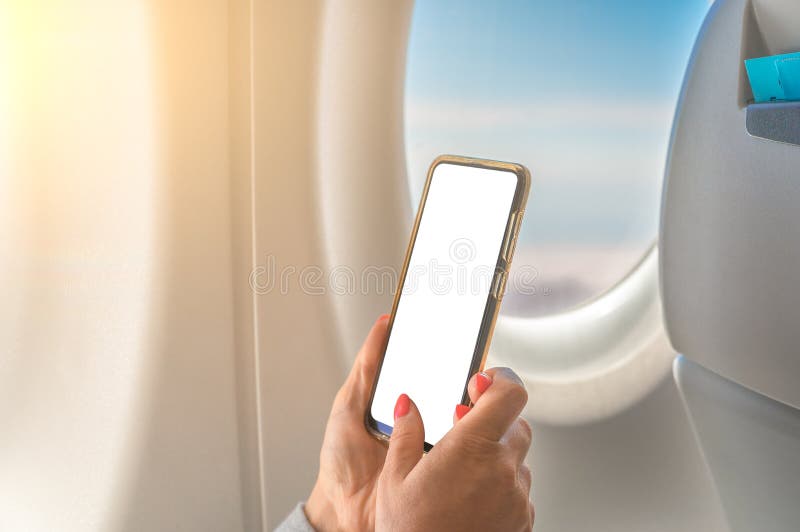Mockup image of a woman holding at smart phone with blank white screen next to an airplane window with clouds and sky background stock photography