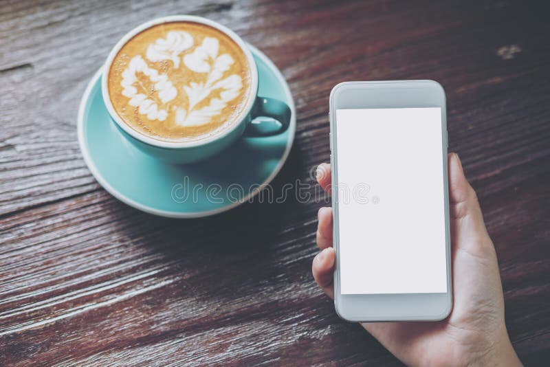 Mockup wizerunek trzyma białego telefon komórkowego z pustym ekranem z błękitną gorącą filiżanką na rocznika drewnianym stole ręk obraz stock