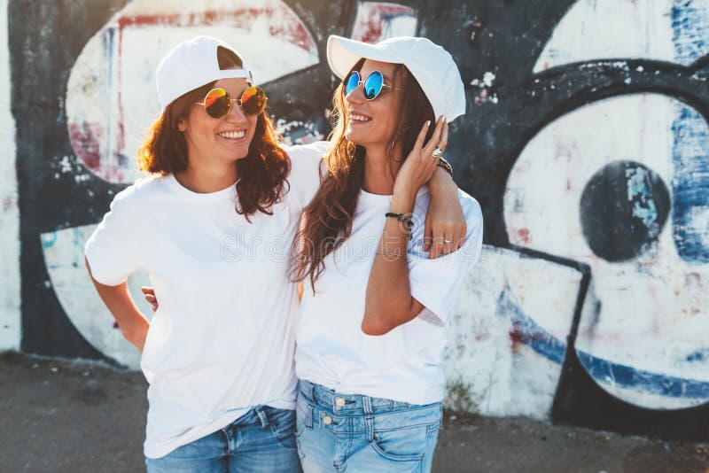 Two models wearing plain white t-shirts and hipster sunglasses posing against street wall. Teen urban clothing style, same look. Mockup for tshirt print store. Two models wearing plain white t-shirts and hipster sunglasses posing against street wall. Teen urban clothing style, same look. Mockup for tshirt print store.