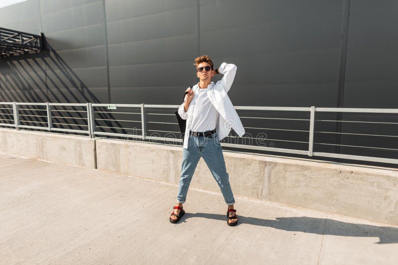 Modello di moda europeo dei pantaloni a vita bassa del giovane in jeans d'annata in una camicia alla moda in sandali alla moda in fotografia stock Modello di moda europeo dei pantaloni a vita bassa del giovane in jeans d'annata in una camicia alla moda in sandali alla moda in fotografia stock