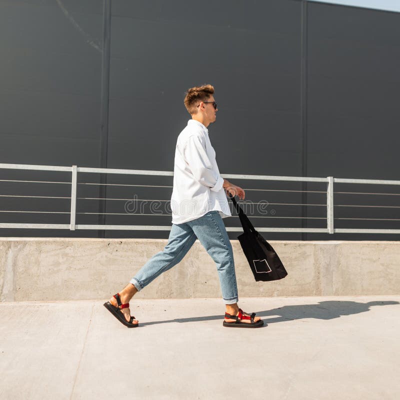 Modello di moda urbano dei pantaloni a vita bassa del giovane in jeans d'annata in una camicia alla moda in sandali alla moda in fotografia stock Modello di moda urbano dei pantaloni a vita bassa del giovane in jeans d'annata in una camicia alla moda in sandali alla moda in fotografia stock