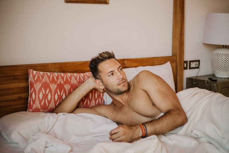 Modello maschio sexy senza camicia che si trova da solo sul suo letto nella sua camera da letto, distogliente lo sguardo con un a fotografia stock