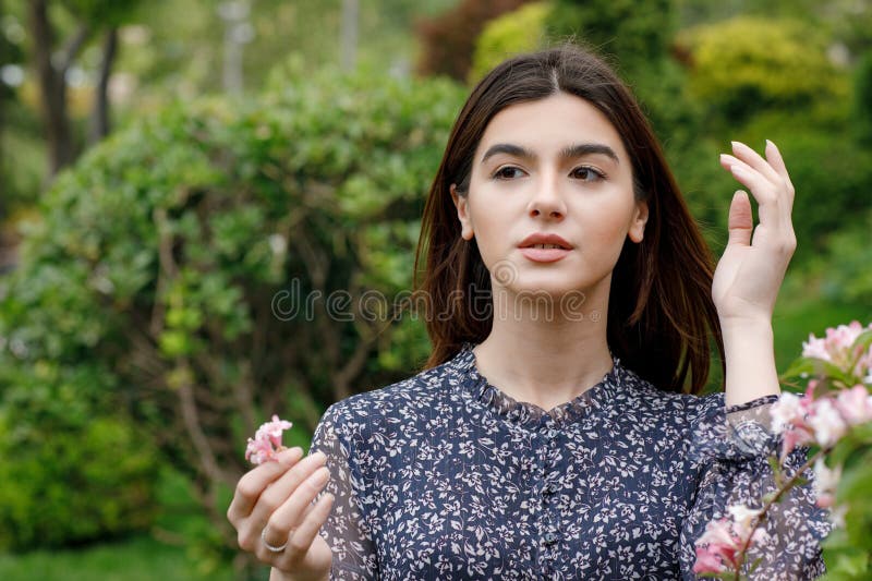 Front view of elegant, brunette model standing in blooming garden, looking forward, holding blossom. Concept of springtime. Front view of elegant, brunette model standing in blooming garden, looking forward, holding blossom. Concept of springtime.