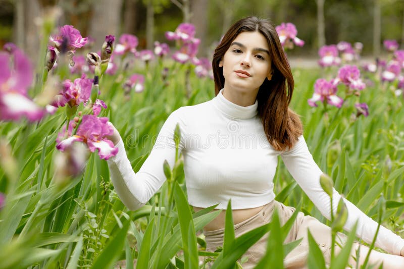 Portrait of elegant brunette model sitting in blooming garden, touching flower by hand, looking at camera. Concept of springtime. Portrait of elegant brunette model sitting in blooming garden, touching flower by hand, looking at camera. Concept of springtime.