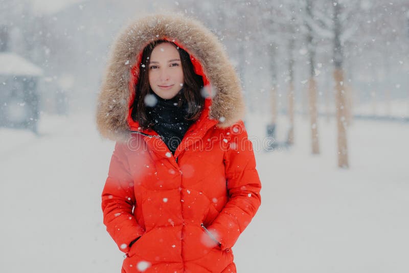 Modelo fêmea bonito no revestimento vermelho, stads exteriores durante a queda de neve, olhares com os olhos escuros na câmera, i fotografia de stock Modelo fêmea bonito no revestimento vermelho, stads exteriores durante a queda de neve, olhares com os olhos escuros na câmera, i fotografia de stock