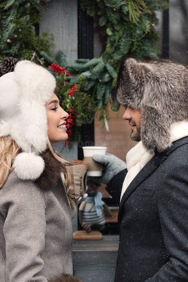 Fashion couple of lovers standing on winter street in snowy weather, man and woman wear fur hat and warm wool coat, smiling, drinking hot drinks. St. Valentines Day, Christmas holidays concept. Fashion couple of lovers standing on winter street in snowy weather, man and woman wear fur hat and warm wool coat, smiling, drinking hot drinks. St. Valentines Day, Christmas holidays concept.