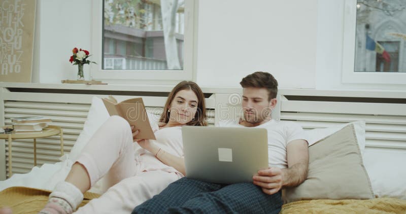 Modern bedroom in the morning closeup young lady reading a book in bed and her partner watching something on the