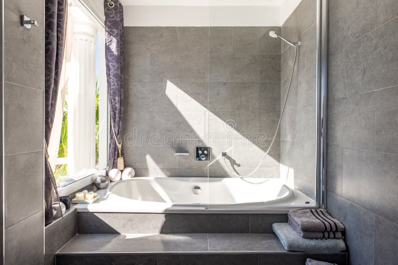 Modern designed grey bathroom with a bath tub and shower head behind a glass with towels and sun shining in from the left royalty free stock photo Modern designed grey bathroom with a bath tub and shower head behind a glass with towels and sun shining in from the left royalty free stock photo