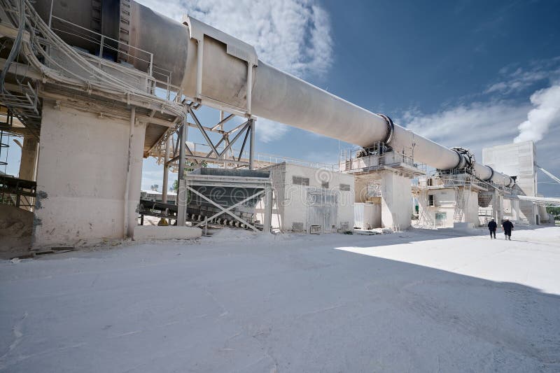 Modern factory of calx production with rotary kiln building under blue sky. Professional workers walk on territory past rotating furnace for limestone roasting