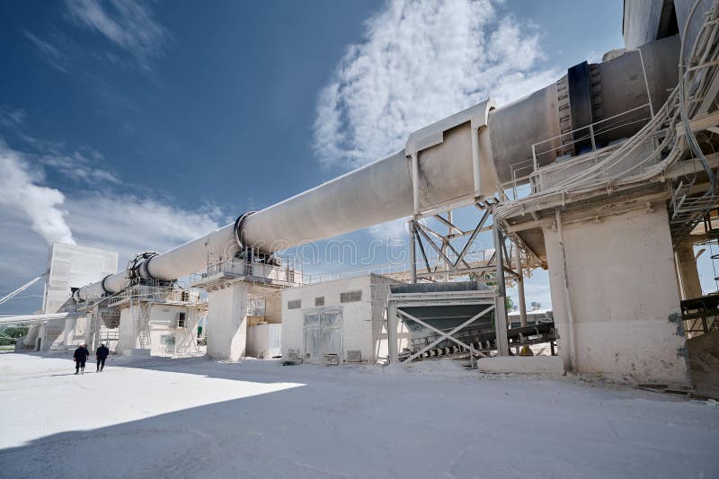 Modern factory of calx production with rotary kiln building under blue sky. Professional workers walk on territory past rotating furnace for limestone roasting