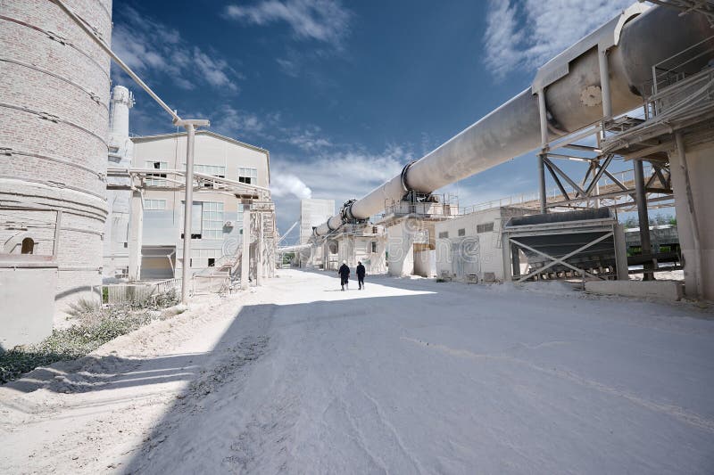 Modern factory of calx production with rotary kiln building under blue sky. Professional workers walk on territory past rotating furnace for limestone roasting