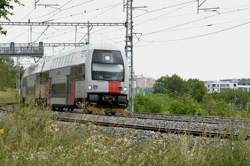 Modern high-speed train moves along railway tracks surrounded by green vegetation. The train has a streamlined design with red and white colors. Overhead electrification wires and pylons indicate an electrified railway system. In the background, there are industrial buildings and lush greenery, suggesting a suburban or semi-urban setting. The sky is overcast, typical of a summer day. Red pylons stock images, royalty-free photos and pictures