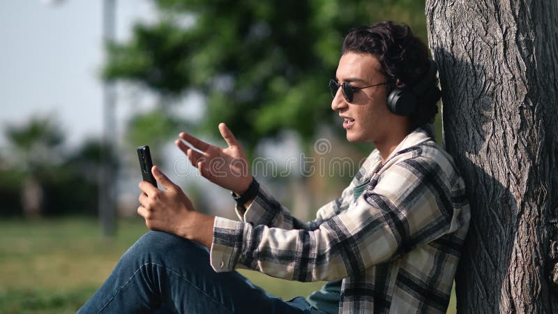 Modern Latino guy in headphones talking online video call use smartphone relaxing at summer park stock footage