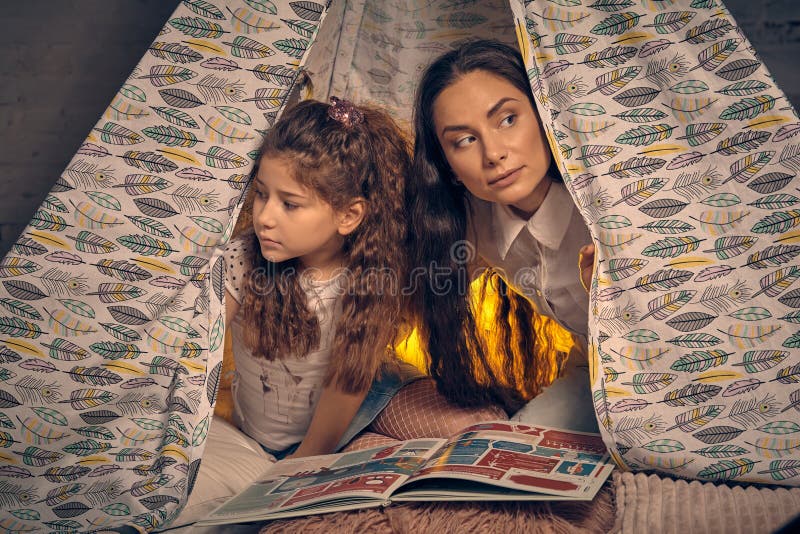 Modern och dottern sitter i ett tipit? lt med n? gra kuddar och l? ser ber? ttelser lycklig familj royaltyfri foto