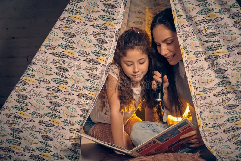 Modern och dottern sitter i ett tipit? lt som l? ser ber? ttelser med ficklampan lycklig familj fotografering för bildbyråer