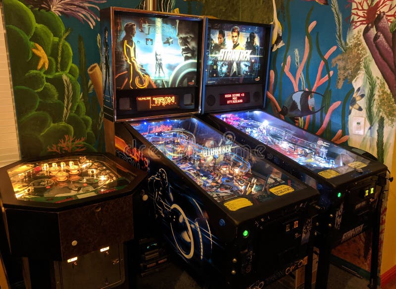 2 modern 2000s, 2010s pinball machines with a 70s cocktail table machine, with an aquatic mural on the wall behind. Eros One, Tron, Star Trek. Tron backglass features Jeff Bridges as Flynn and Clu, Garrett Hedlund as Sam, and Olivia Wilde as Quorra. Star Trek backglass features Chris Pine as Captain James Kirk, Zoe Saldana as Uhura, Zachary Quinto as Spock, Eric Bana as Nero, and Benedict Cumberbatch as Khan. 2 modern 2000s, 2010s pinball machines with a 70s cocktail table machine, with an aquatic mural on the wall behind. Eros One, Tron, Star Trek. Tron backglass features Jeff Bridges as Flynn and Clu, Garrett Hedlund as Sam, and Olivia Wilde as Quorra. Star Trek backglass features Chris Pine as Captain James Kirk, Zoe Saldana as Uhura, Zachary Quinto as Spock, Eric Bana as Nero, and Benedict Cumberbatch as Khan.
