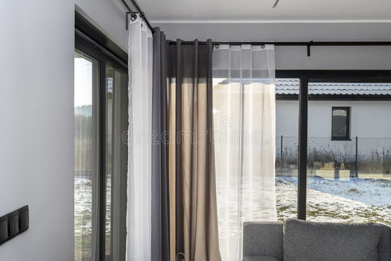 A modern and simple living room in the house with large terrace windows with curtains and a gray sofa.