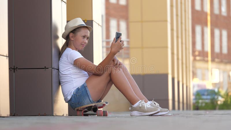 Modern tonåring som sitter på en skateboard på gatan med en smartphone som pratar online. moderiktig tjej läser