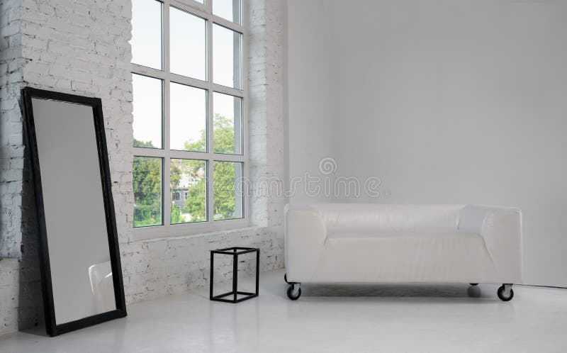 Modern white sofa and large black framed mirror in white minimalistic room with big window